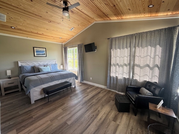 A bedroom with a wooden ceiling, a bed with a headboard, a nightstand, a bench, a chair, a TV, and a window with curtains.