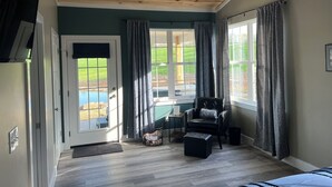 A room with a wooden ceiling, a door leading outside, a black leather chair, a small ottoman, and a window with curtains. - Private Cabin on 90 Acres! (Troy)