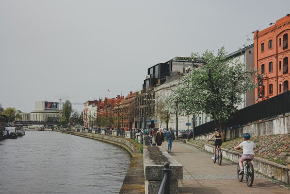 A riverside walkway with buildings, a bridge, and cyclists.