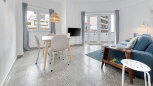 Una sala de estar moderna con una mesa de comedor, sillas, un sofá, un televisor y un balcón con vistas a edificios.