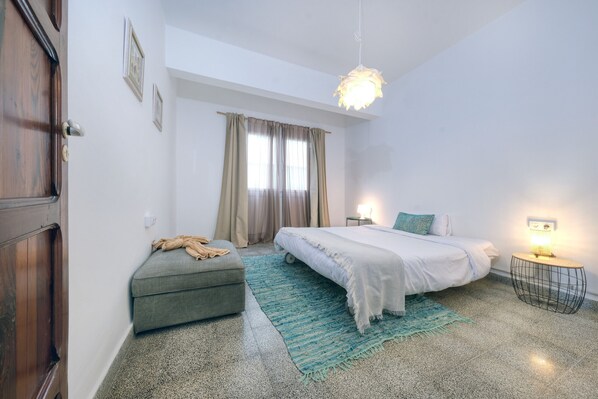 A bedroom with a bed, a small seating area, a window with curtains, and a hanging light fixture.