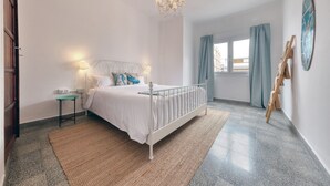 Un dormitorio con cama blanca, una mesita de noche pequeña, una lámpara de araña, una ventana con cortinas y una silla de madera junto a la pared.