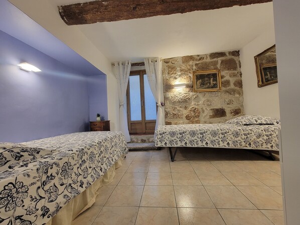 A room with two beds, a stone wall, a wooden ceiling beam, and a window with curtains.