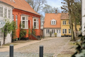 A cobblestone street lined with traditional European buildings, featuring a mix of red and yellow facades, and a visible sign on the left.
