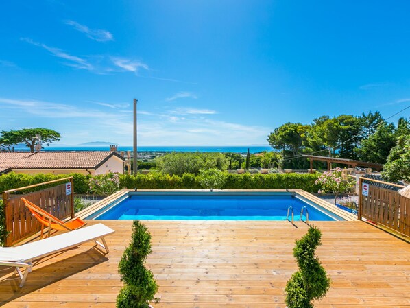 A wooden deck with a swimming pool, lounge chairs, and a view of the sea.