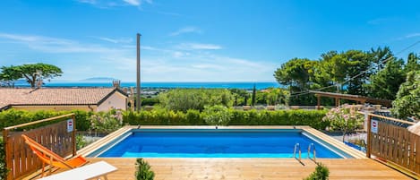 A wooden deck with a swimming pool, lounge chairs, and a view of the sea.