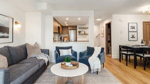 Una sala de estar moderna con un sofá gris, dos sillones azules, una mesa de centro y un comedor con una mesa y sillas de madera.