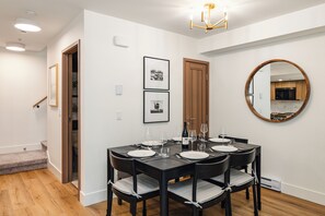 Un comedor con una mesa de madera oscura dispuesta para cuatro, platos blancos y copas de vino. Hay dos fotografías enmarcadas en la pared, una puerta de madera y un espejo redondo que refleja la cocina.
