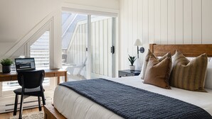 Un dormitorio con cama, escritorio con computadora portátil, silla, mesita de noche con lámpara y vista al exterior a través de una puerta corrediza.