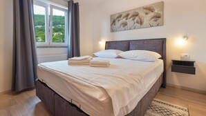Un lit bien fait, avec du linge de lit et des serviettes blanches, une tête de lit grise et une vue sur la verdure par une fenêtre ornée de rideaux gris.