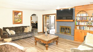 Una sala de estar con sofá, sillón y una mesita de centro. Hay una chimenea con un televisor encima y una estantería llena de libros.