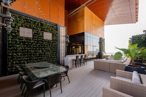 An outdoor dining area with a glass table and chairs, a bar with stools, and a seating area with cushions.