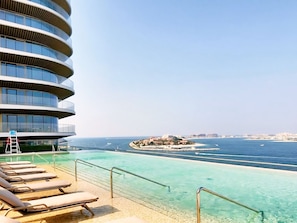 Un hotel moderno con fachada curva, zona de piscina con agua cristalina y vistas al mar y edificios lejanos.