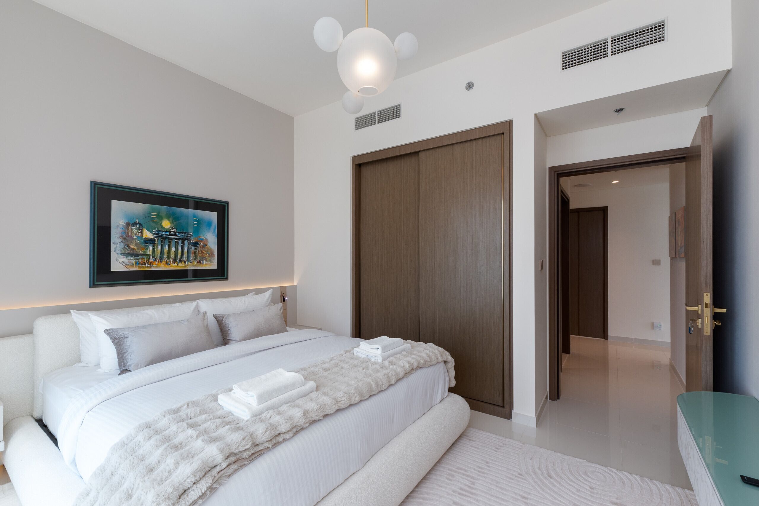 Une chambre d’hôtel moderne avec un grand lit, un tableau représentant un paysage urbain encadré, une armoire et une salle de bain dotée d’un plan de toilette vert.