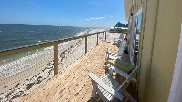 Una terraza de madera con barandilla que da a una playa, dos sillas Adirondack y un edificio con ventana.