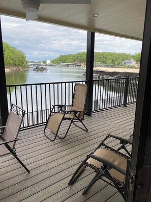 Una terraza de madera con dos sillas y una mesa, con vistas a un río y edificios con vegetación al fondo.