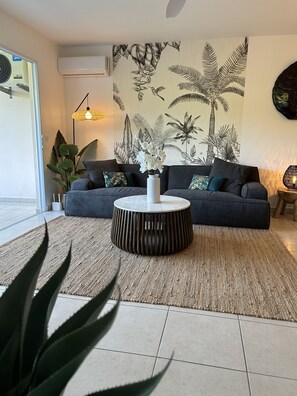 Een woonkamer met een grijze bank, een ronde salontafel en een muur met een tropisch bladmotief.