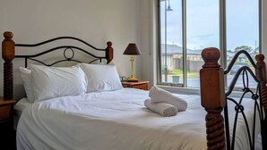 A neatly made bed with white linens, a wooden headboard, and a window with a view of a residential area. - Spacious home near Bellarine Peninsula Trail (Curlewis)