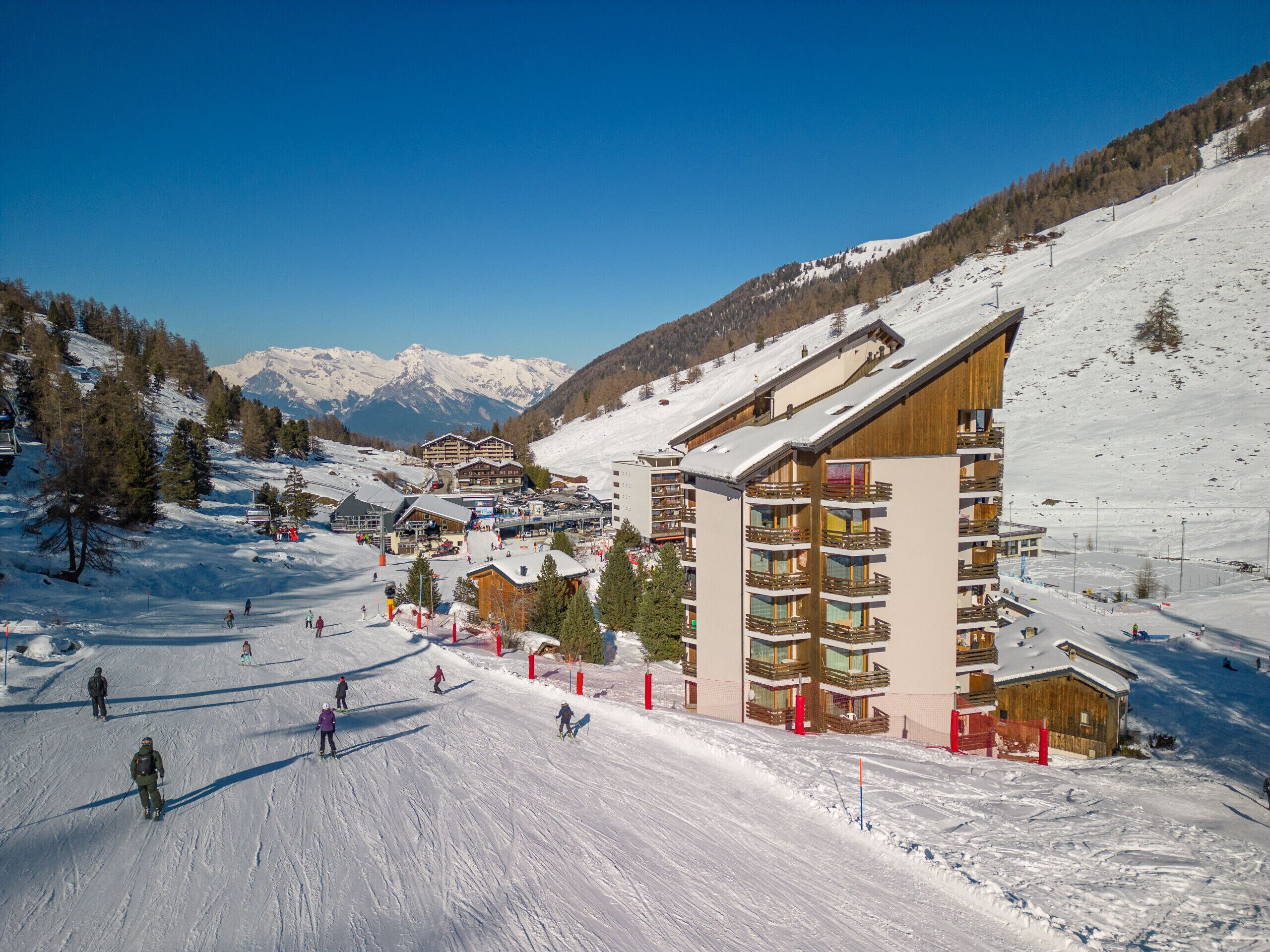Une station de ski comprenant plusieurs bâtiments, dont un imposant édifice à étages doté de balcons, entouré de pentes enneigées et avec des montagnes en toile de fond.