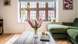 A living room with a green sofa, a wooden coffee table with a vase of flowers, and a window with a view of buildings. - 57A Thoroughfare, Woodbridge (Woodbridge)