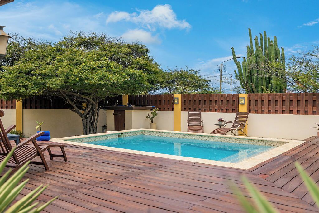 Una piscina rectangular con muelle de madera, rodeada por una cerca de madera y un árbol.