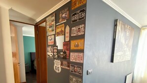 Un couloir aux murs bleu foncé, orné de diverses plaques d’immatriculation anciennes. Le sol est carrelé, et le couloir mène à une pièce où se trouvent un lit et un bureau.