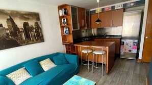Un espace de vie compact comprenant un canapé de couleur turquoise, une petite table basse, un coin cuisine avec un réfrigérateur et un tableau représentant un paysage urbain accroché au mur.