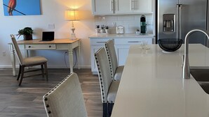 Una cocina moderna con armarios blancos, un refrigerador de acero inoxidable, una mesa de comedor con sillas y una computadora portátil sobre un escritorio.