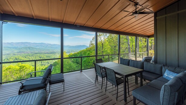 A covered outdoor area with a table and seating, large windows offering a view of green mountains.