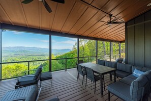 A covered outdoor area with a table and seating, large windows offering a view of green mountains. - Bear Ridge Retreat | Pet-Friendly New Build with VIEWS! (Old Fort)