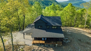 A two-story cabin with a dark roof, surrounded by a forested area with a view of distant hills.