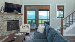 A living room with a stone fireplace, a flat-screen TV, a sofa, a coffee table, and a view of mountains through a glass door.