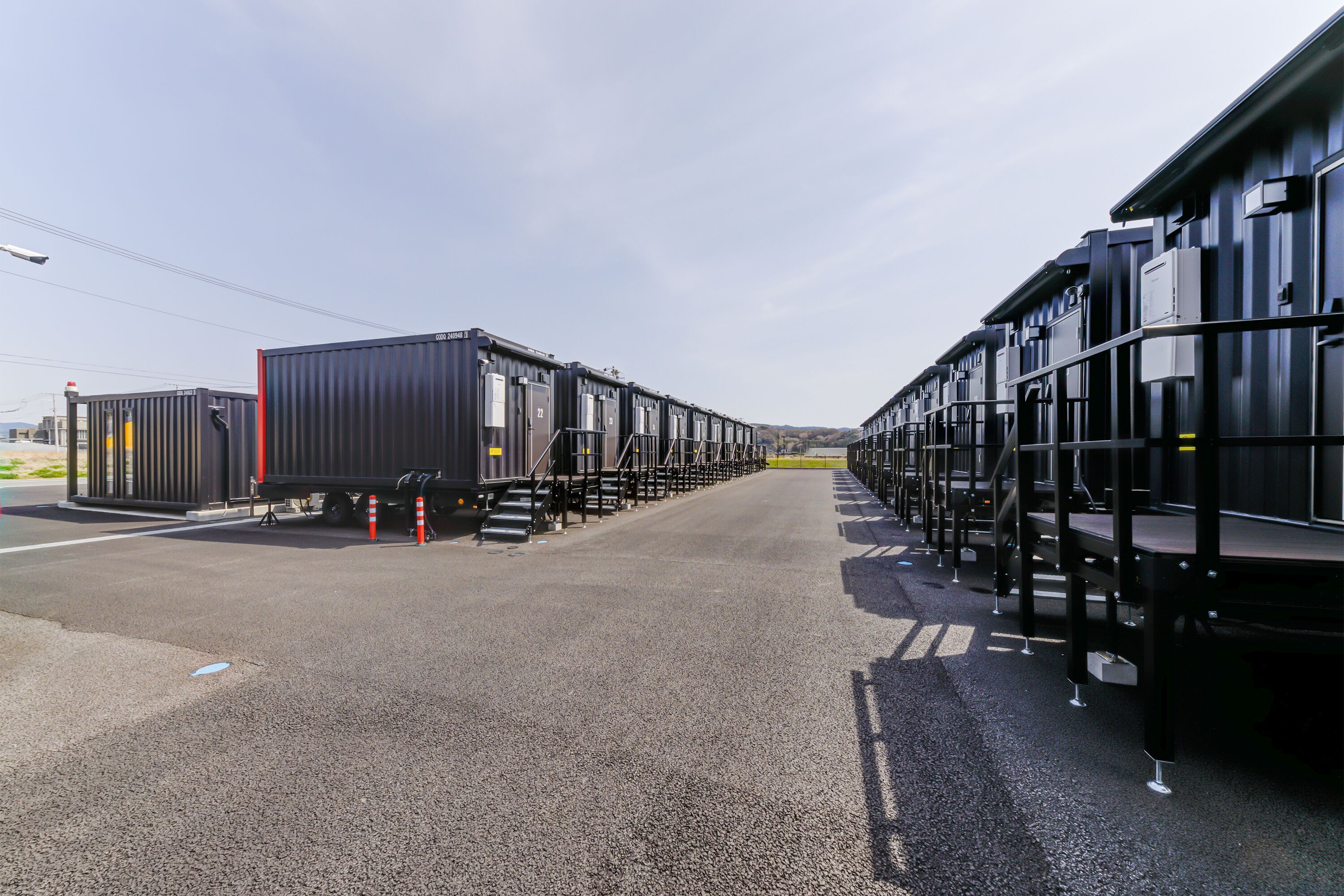 A row of shipping containers arranged in a linear formation, with a clear sky overhead and a paved surface in the foreground.