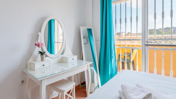 A hotel room with a glass desk, a bed with white linens, a large window with a view of a balcony, and a mirror.
