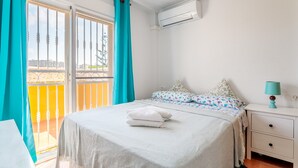 A bedroom with a bed, a nightstand, a lamp, a window with curtains, and an air conditioning unit.