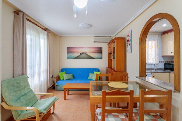 A living room with a sofa, a dining table with chairs, a wooden cabinet, and a kitchen visible through an archway. - Apartment 'Pretty Bungalow' with Mountain View, Wi-Fi and Air Conditioning (Santa Pola)