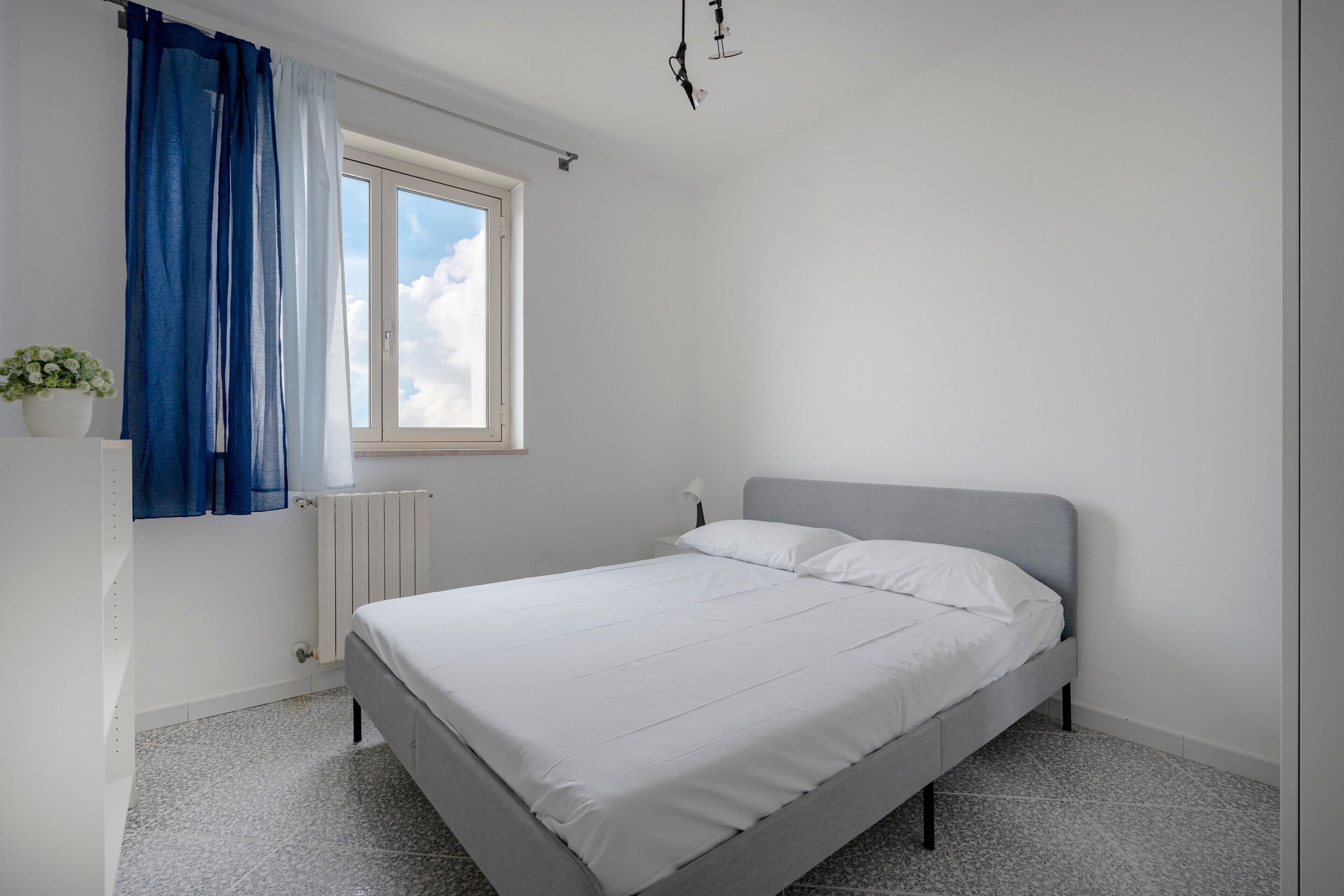 Ein Schlafzimmer mit einem Bett, einem Fenster mit Vorhängen, einem Heizkörper und einer Topfpflanze.