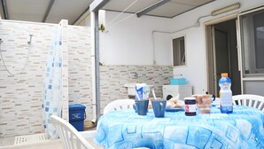 A covered outdoor dining area with a table set for two, a shower, and a white tiled wall.
