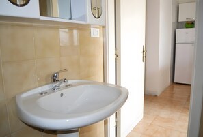 Un baño con lavamanos blanco, un espejo y una puerta que da a una zona de cocina.