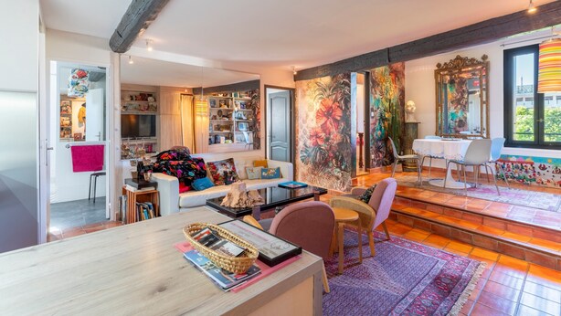 Een goed verlichte woonkamer met een bank, een eettafel en stoelen. Er is een keuken met kasten en een eettafel. De kamer heeft een groot raam en een deur die naar een andere ruimte leidt.