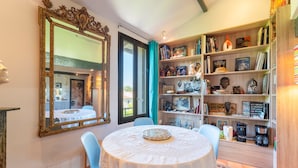 Een eetgedeelte met een ronde tafel, stoelen, een grote, sierlijke spiegel en een boekenplank vol boeken en decoratieve objecten.