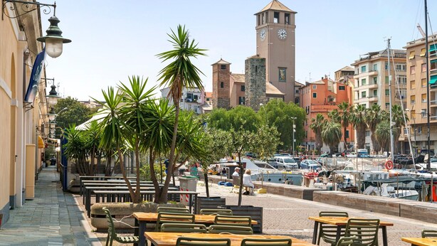 A coastal town with a prominent clock tower, outdoor seating, and a marina. - Cruise Apartment with Private Terrace, Balcony, and Wi-Fi (Savona)