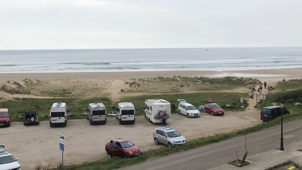 A beachside parking area with RVs and cars, a road with a sidewalk, and a beach in the background.