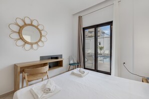 A hotel room with a bed, a desk, a chair, a TV, and a large window with a view of buildings and a pool.
