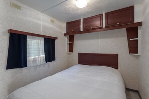A bedroom with a bed, a window with curtains, built-in wooden cabinets, and a ceiling light. - Mobile Home at Camping Le Front De Mer – Excludes Camping Fees – Shared Pool & Air Conditioning (Argelès-sur-Mer)