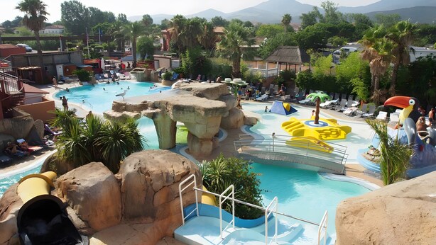 Une grande piscine extérieure, agrémentée de rochers et de toboggans, entourée de palmiers et bordée par un paysage montagneux.
