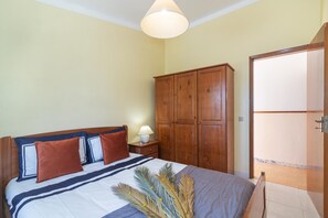 Ein Schlafzimmer mit Bett, Holzschrank, Nachttisch, Lampe und Blick durch eine offene Tür.