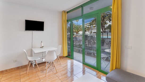 A room with a TV, a desk, chairs, a bench, and large windows with yellow curtains.