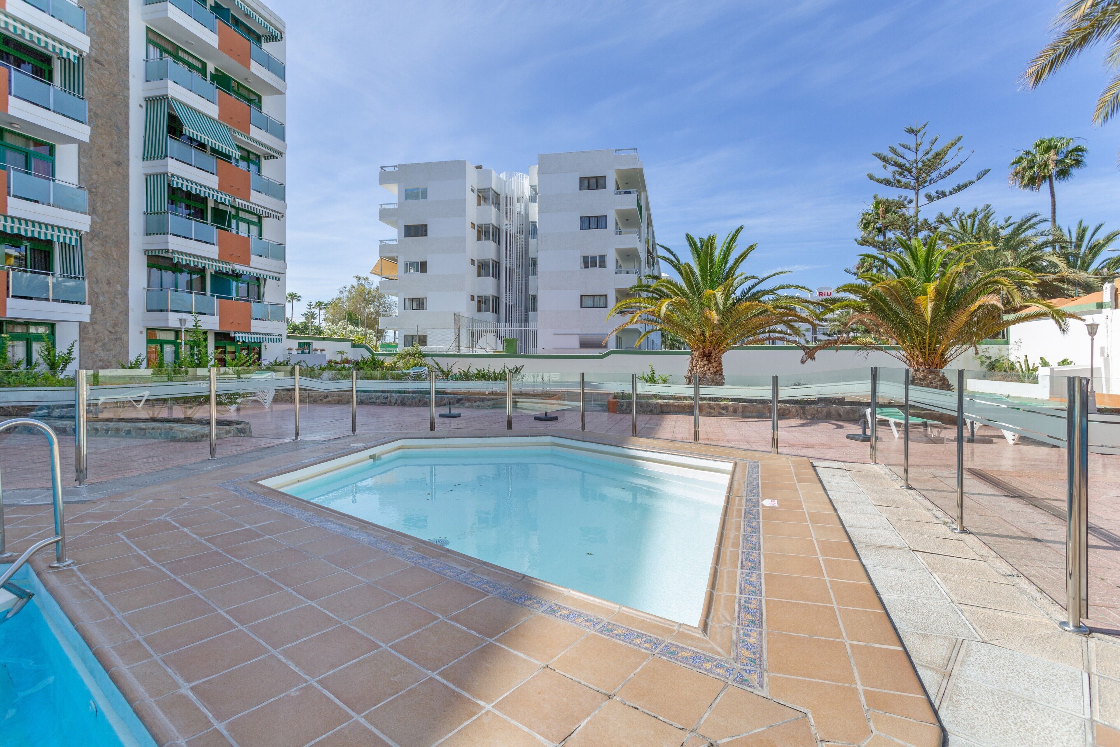 A swimming pool with a tiled deck, surrounded by palm trees and a multi-story building with balconies.