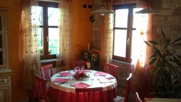 A round dining table with red chairs, set for four, in a room with stone walls and large windows with floral curtains.